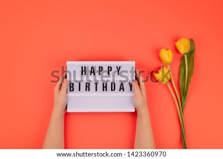 Happy birthday flat lay. Children hands holding light box with text Happy birthday and bouquet of yellow tulips on coral background. 