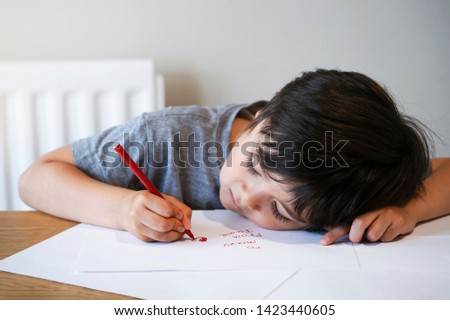 Emotional portrait of child writing a messages to his mother, Preschool kid using red colour writing and drawing on white paper, Little boy lying head down on table drawing red heart for mam. 