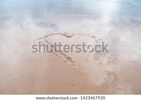 minimalist image of a large heart drawn in the sand on a flat beach with gentle tide going out and blue sky reflecting in remaining water
