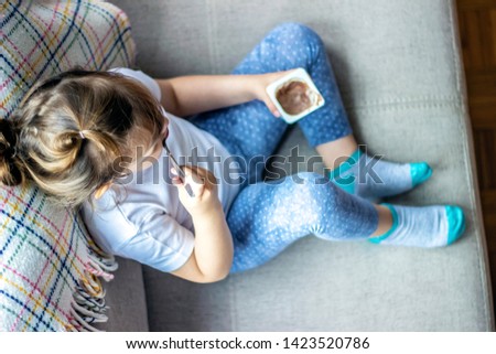 Top view of the cute girl eating her favorite chocolate dessert. Beautiful little girl enjoying her moment. Toddler in white tshirt sitting on bed alone and eating afternoon snack. 