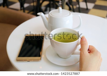 Woman's Hand Holding Hot Green Tea Cup on the Round Table with Teapot and Smartphone