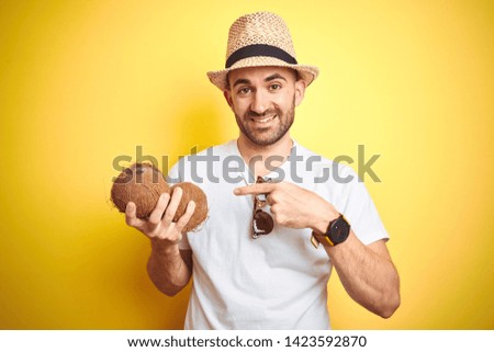 Young man wearing summer hat and holding coconut fruit over yellow background very happy pointing with hand and finger