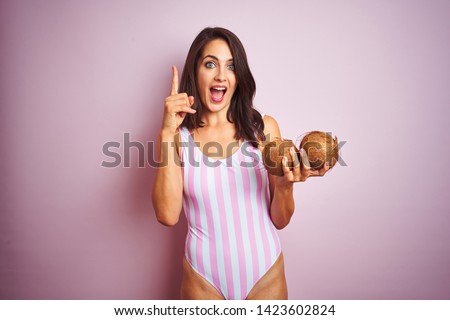 Beautiful woman wearing pink striped swimsuit holding coconuts over isolated pink background surprised with an idea or question pointing finger with happy face, number one