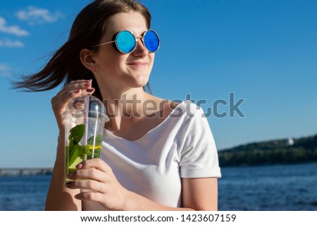 young happy girl sits in blue glasses against the background of the sea and the sky holds a mojito and smiles, she is resting on the beach and looking into the distance