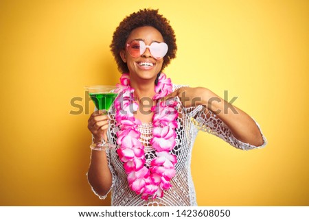 Young african american woman with afro hair wearing flower hawaiian lei and drinking a cocktail with surprise face pointing finger to himself