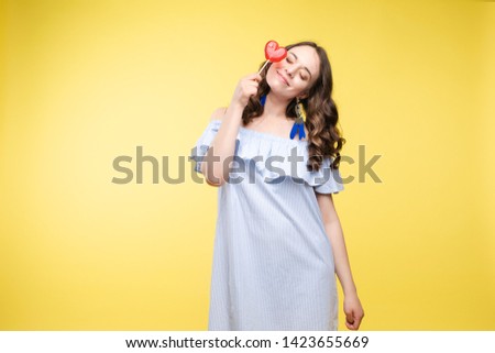 Beautiful and seductive woman wearing like sweet doll, posing at studio with candy on stick. Confident pretty woman in dress andsunglasses, holding hand on waist. Fashion, glamour.