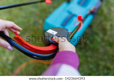 Women's hands on the handle of an electric lawn mower. Red start button for lawn mower. The girl includes a lawn mower and starts mowing the lawn.