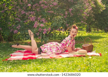sweetheart charming pinup girl in a summer dress on a checkered blanket in the Park near the bushes of lilac enjoys life and leisure alone. pretty happy caucasian woman on the grass picnic.