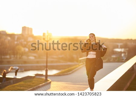 Young happy woman with a good mood in sunglasses in a stylish t-shirt in the city on the background of a bright orange sunset. Joyful girl model enjoys the sun.