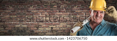 Male Contractor In Hard Hat Holding Construction Plans In Front Of Old Brick Wall Banner with Copy Space.