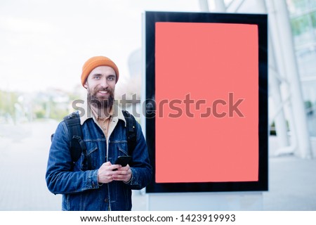 Hipster beard caucasian man in a unformal wear using cell phone while standing outdoors next to the white city advert coral banner mock-up; man entrepreneur near template of a blank vertical billboard