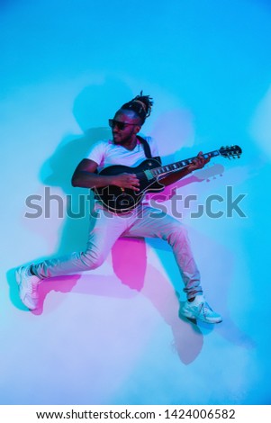 Young african-american musician playing the guitar like a rockstar on blue studio background in neon light. Concept of music, hobby. Joyful attractive guy improvising. Retro colorful portrait.