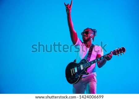 Young african-american musician playing the guitar like a rockstar on blue studio background in neon light. Concept of music, hobby. Joyful attractive guy improvising. Retro colorful portrait. Royalty-Free Stock Photo #1424006699