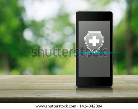 Cross shape with shield flat icon on modern smart mobile phone screen on wooden table over blur green tree in park, Business healthy and medical care insurance online concept
