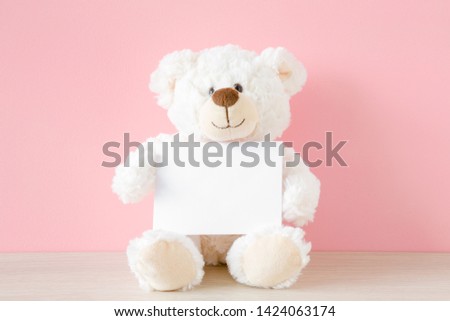 Smiling teddy bear sitting at pastel pink wall and holding white card. Empty place for inspiration, emotional, sentimental text, lovely quote or sayings for good mood. Front view.