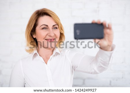 portrait of cheerful mature business woman taking selfie with smart phone over white brick wall