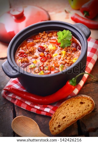 Hot and spicy fresh made Mexican chili still in an iron pot, with a raw chili-pepper and a slice of bread