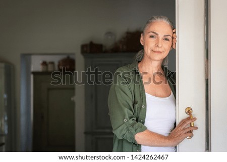 Portrait of senior woman leaning against door at home and looking at camera. Smiling mature woman standing at doorway and looking at camera. Portrait of old lady relaxing and thinking with copy space. Royalty-Free Stock Photo #1424262467