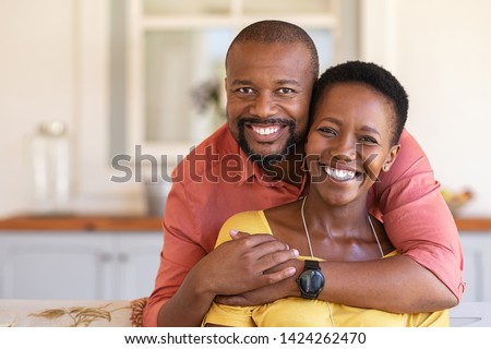 Happy mature black couple bonding to each other and smiling while sitting on couch. Portrait of smiling black man embrace his wife from behind and looking at camera. Royalty-Free Stock Photo #1424262470