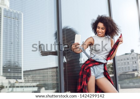 Young woman in the city street standing near window taking selfie pistures on smartphone looking camera smiling cheerful