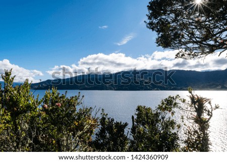 The panorama of lake surrounded of mountains, trees, nature. The sun has rays light that illuminates the photography.  Clouds on mountains.