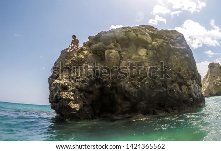 A young man sitting on a boulder protruding from the sea. Sharp surface of the rock looks dangerous to climb on. Sea below the rock is calm, and in the turquoise shade. Beautiful site in Cyprus.