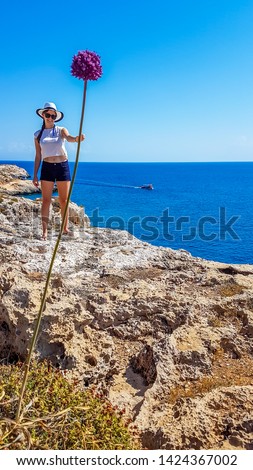 A girl holding a gigantic flower in her hand. Changing the perspective, fun photography. Girl is wearing a hat and shorts. Behind there is an open sea. Barren landscape of Cape Greco, Cyprus.