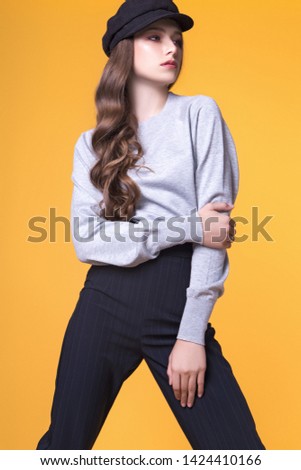 Beautiful teen girl with long hair posing in the studio on a yellow background, wearing a gray sweater, black pants and a cap. Fashion concept