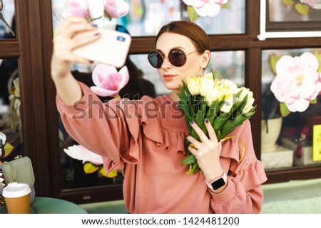 girl in a pink sweater, sitting on a summer playground near a cafe and taking a selfie with tulips