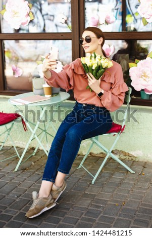 girl in a pink sweater, sitting on a summer playground near a cafe and taking a selfie with tulips