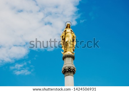Golden religious statue, illuminated by the sun, of the Virgin Mary on top of a pedestal, with a background of blue sky and clouds.