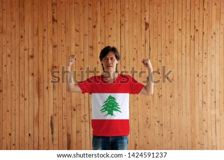 Man wearing Lebanon flag color of shirt and standing with raised both fist on the wooden wall background, triband of red and white, charged with a green Lebanon Cedar.