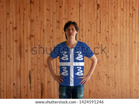 Man wearing Martinique flag color shirt and standing with akimbo on the wooden wall background, four white snake on blue field and white cross in the center.