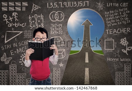 Close-up of a boy reading book with chalkboard and success road via keyhole