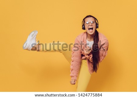 Young cheerful brunette in glasses dancing, listening music in headphones high lifting her leg dressed in yellow pants, white shoes, pink sweater and headphones on her head over yellow background.  Royalty-Free Stock Photo #1424775884