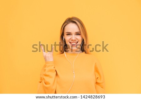 Happy blonde in an orange sweatshirt and headphones is on a yellow background, listens to music and shows a sign of heavy metal, looks into the camera and smiles. Isolated