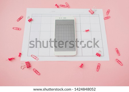 White smartphone lies on a paper calendar. Pink background. Bright pink clips and pins are scattered around.