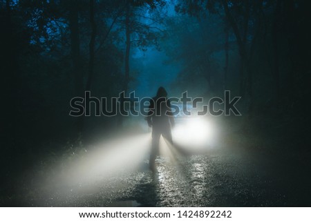 A sinister hooded figure standing in front of a car. On a spooky forest road on a foggy evening. Royalty-Free Stock Photo #1424892242