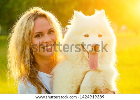beautiful curly blonde smiling toothy happy girl in denim shorts are sitting at glass hugging white fluffy cute samoyed dog in the summer park sunset rays field background . pet and hostess