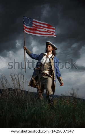 American revolution war soldier with flag of colonies over dramatic landscape. 4 july independence day of USA concept photo composition: soldier and flag. Royalty-Free Stock Photo #1425007349