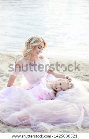 Mother and her little girl enjoying lake view and relaxing on the beach on a sunny day in beautiful dresses. A cheerful child and her mother are enjoying the sunny morning near the water. family love 