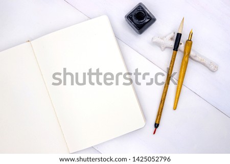 Blank page of sketchbook with calligraphic brush, nib and black ink jar. Minimal flat lay with writing or drawing tools. Oriental mockup for calligraphy or ink sketching. Artist work desk composition