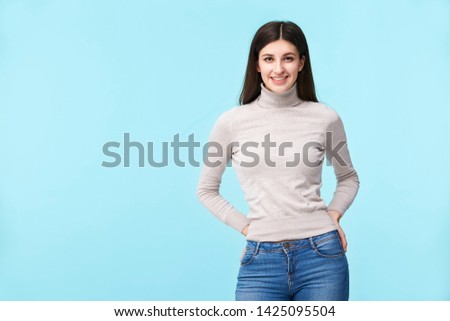 portrait of a beautiful young caucasian woman, standing, hands on hips, looking at camera smiling, isolated on blue background Royalty-Free Stock Photo #1425095504