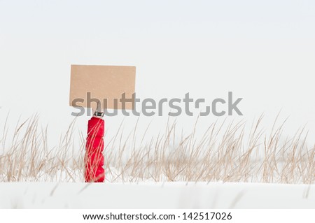 Arm in warm glove and red jacket holding blank board sticking out of snow. Snow and stubble in foreground, winter field and sky in background. Creative commercial or signpost.