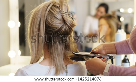 Hairtician twirls curls with flat iron hair. Back view. Hairdressing services. Creation of evening hairstyles fashionable stylish women's hairstyles. Hair styling process. Courses in hairdressing. Royalty-Free Stock Photo #1425214076