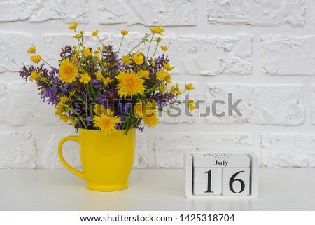 Wooden cubes calendar July 16 and yellow cup with bright colored flowers against white brick wall. Template calendar date for your design Copy space.