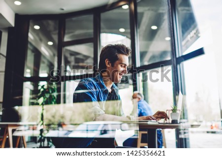 Side view of cheerful millennial hipster guy in casual wear feeling good during online chatting with followers at web blog connected to public wifi in cafeteria, happy man searching jokes on website