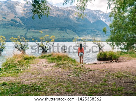 Woman walking to row of Willow Trees, on lake in Glenorchy. Beautiful landscape, with blue lake, mountains and clear sky. Nature, travel, tourism, outdoor concepts. Shot near Queenstown, New Zealand  