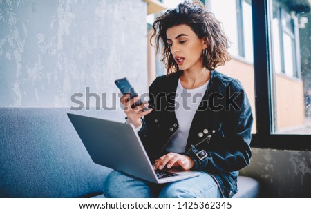 Attractive young woman with short haircut reading received notification on smartphone while updating software on laptop sitting on couch in coworking.Charming female freelancer sending sms on cellular