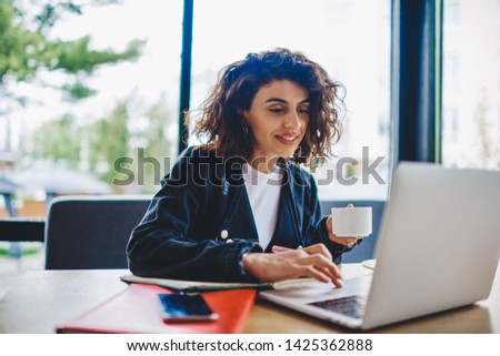 Smiling young woman with short haircut holding coffee cup and reading news on website sitting at laptop in cafe interior.Casual dressed hipster girl enjoying caffeine beverage updating profile in app